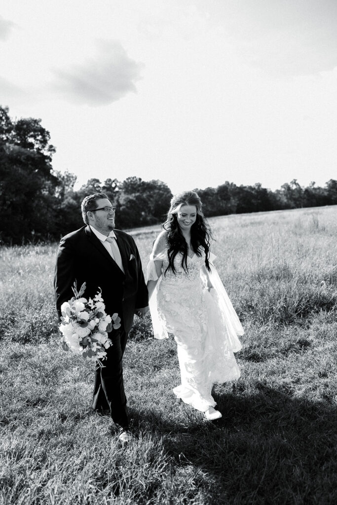 Black and white image of a smiling couple walking hand in hand through a large field on their micro-wedding day at Lost Hill Lake Events in St. Clair, Missouri.
